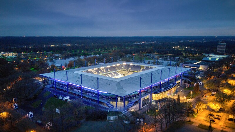 Eine Luftaufnahme zeigt das Fußballstadion in Duisburg bei Dämmerung. Das Stadion leuchtet blau.