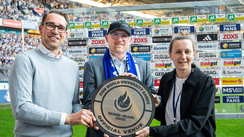 Zwei Männer und eine Frau nehmen vor einer gläsernen Sponsorenwand mit Logos auf einem Rasen in einem Stadion eine Plakette entgegen.