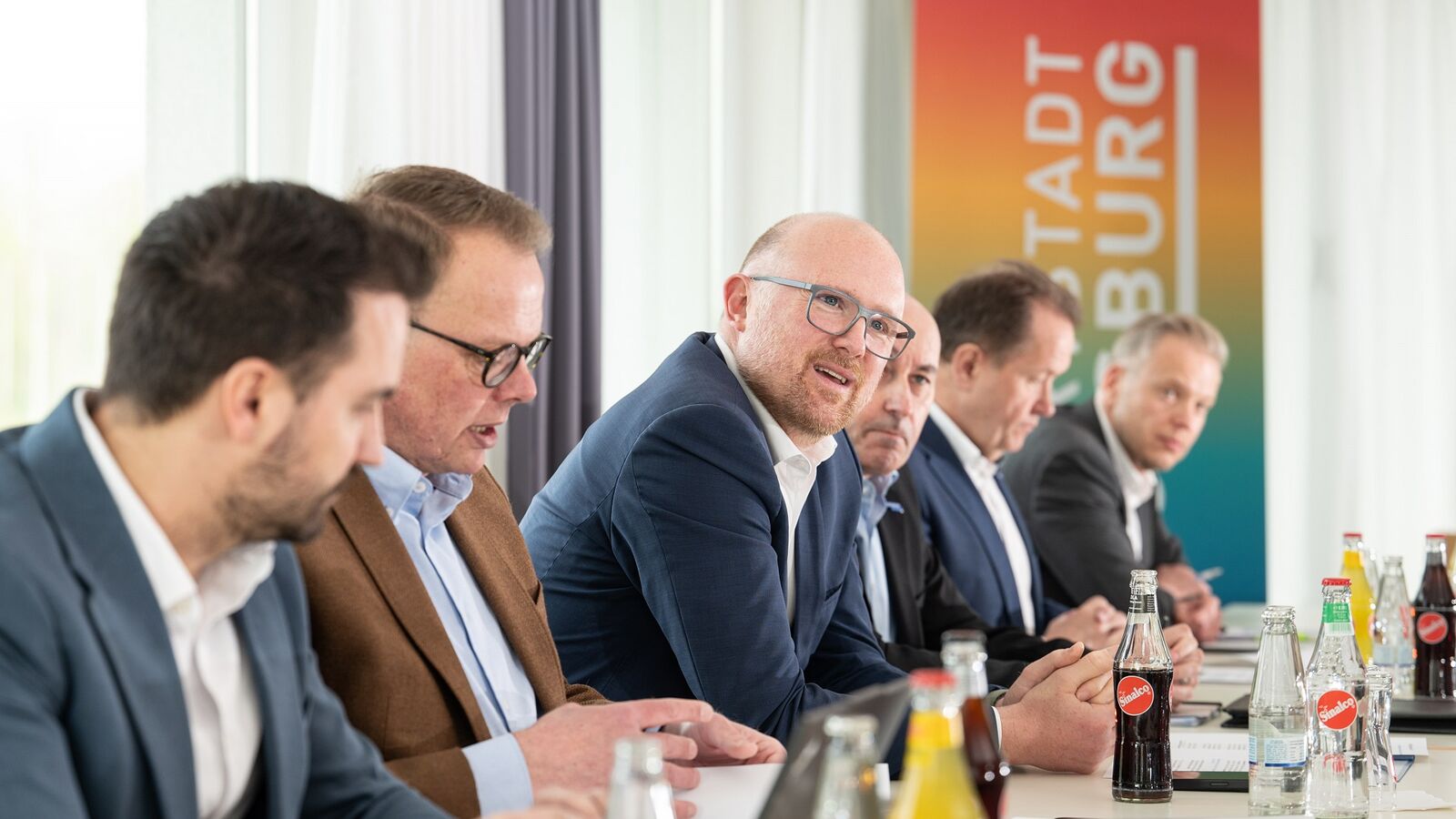 Sechs Männer in Anzügen sitzen nebeneinander an einem Tisch. Auf dem Tisch befinden sich mehrere Getränke und Papiere. Einer der Männer ist am sprechen.